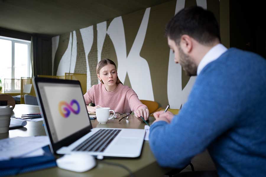 A woman in a pink sweater and a man in a blue sweater are sitting at a table, discussing work. A laptop with a colorful graphic is in the foreground.
